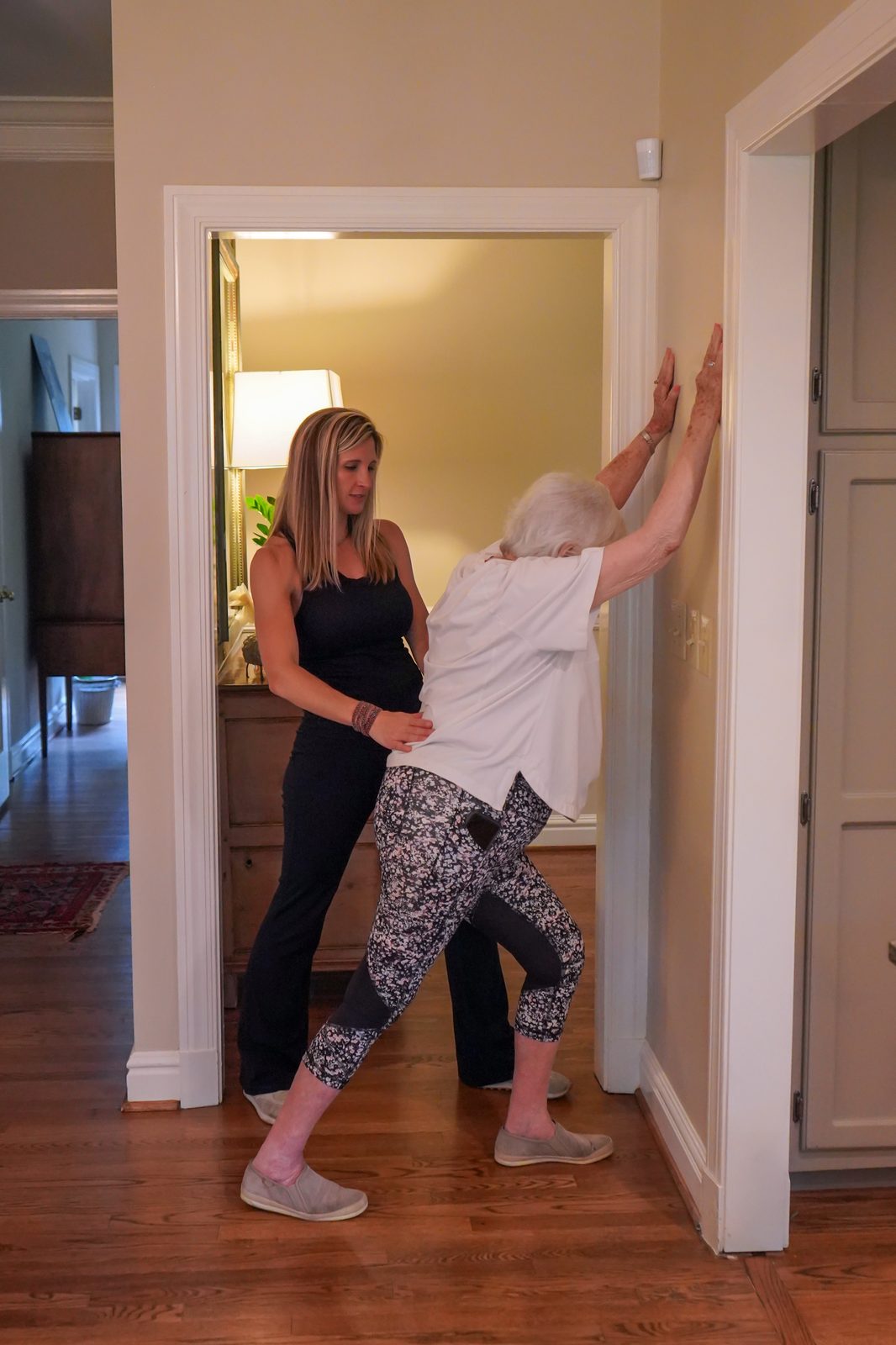 Trainer guiding a senior woman through a supported standing calf stretch against a wall