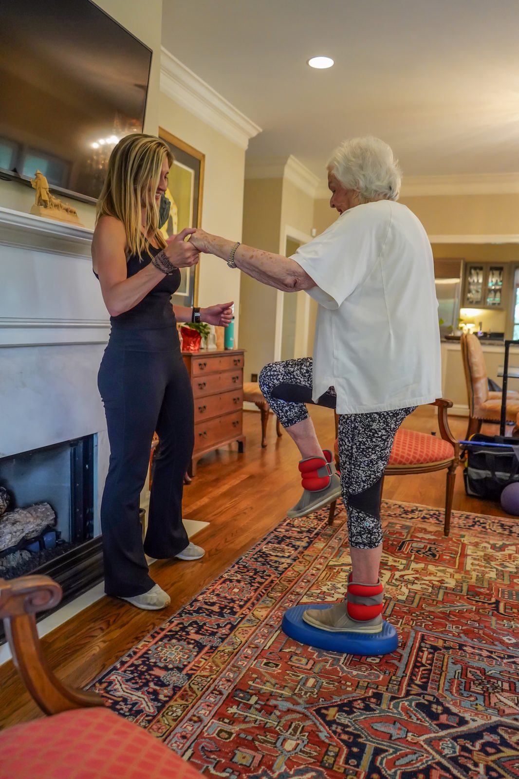 Senior woman balancing on a stability pad with ankle weights while the trainer steadies her hands