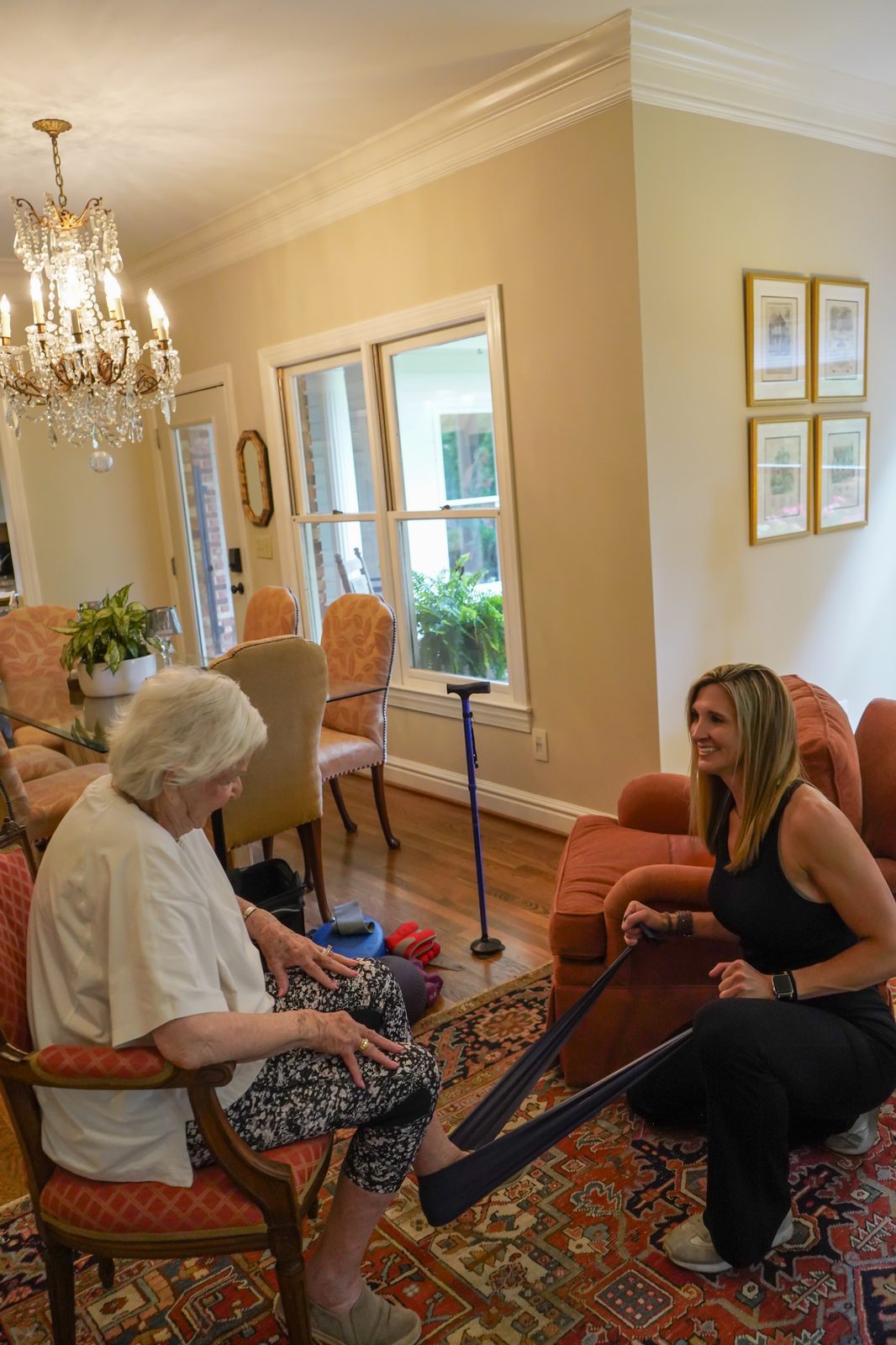 Graceful Aging trainer assisting a senior woman with a seated overhead dumbbell exercise at home