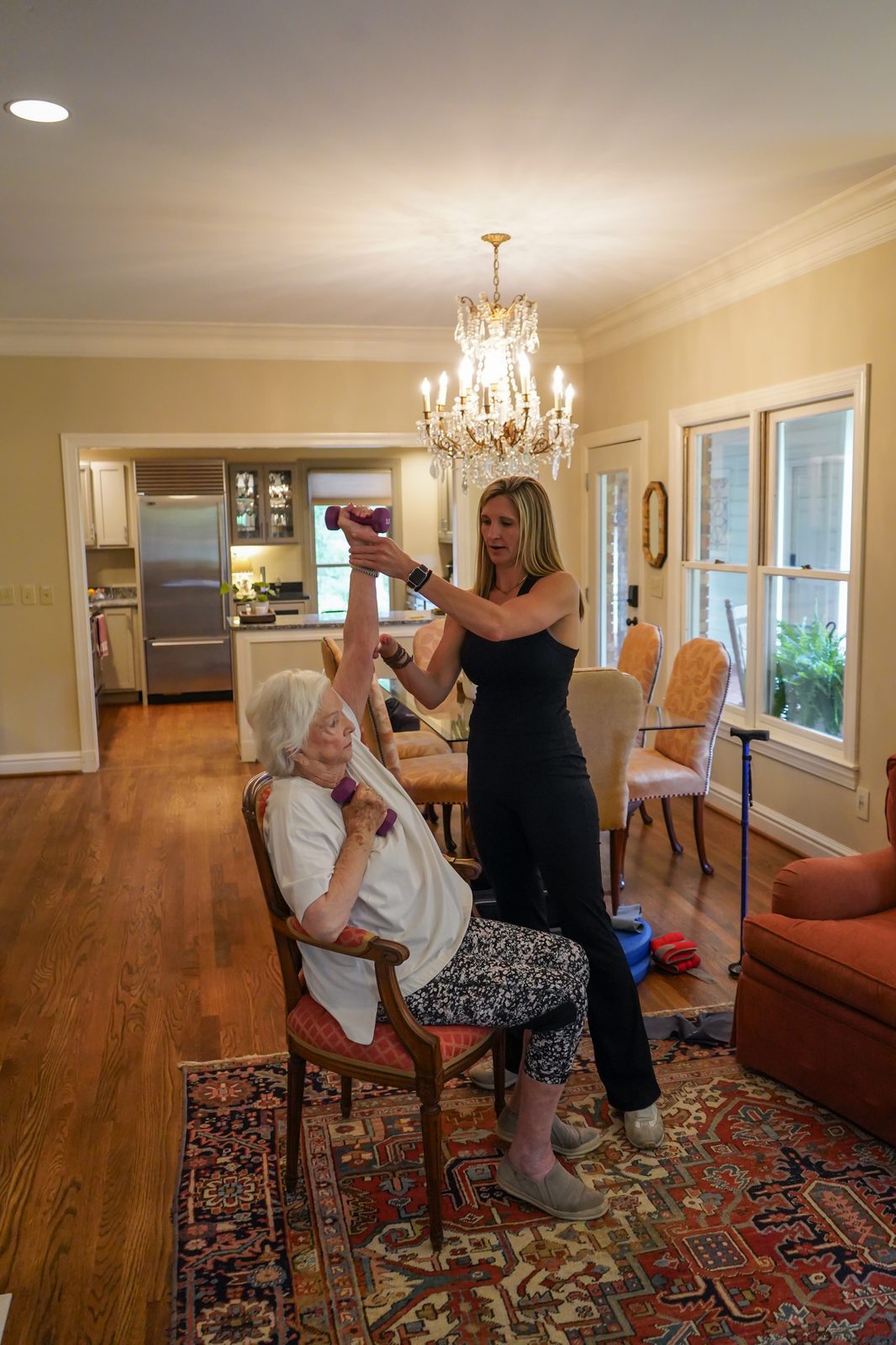 A Graceful Aging trainer guiding a senior woman through a seated resistance-band leg exercise in her Birmingham home