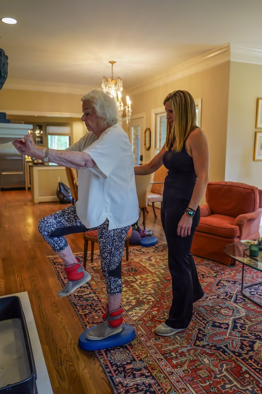Senior woman doing a seated overhead dumbbell press with trainer assistance at home